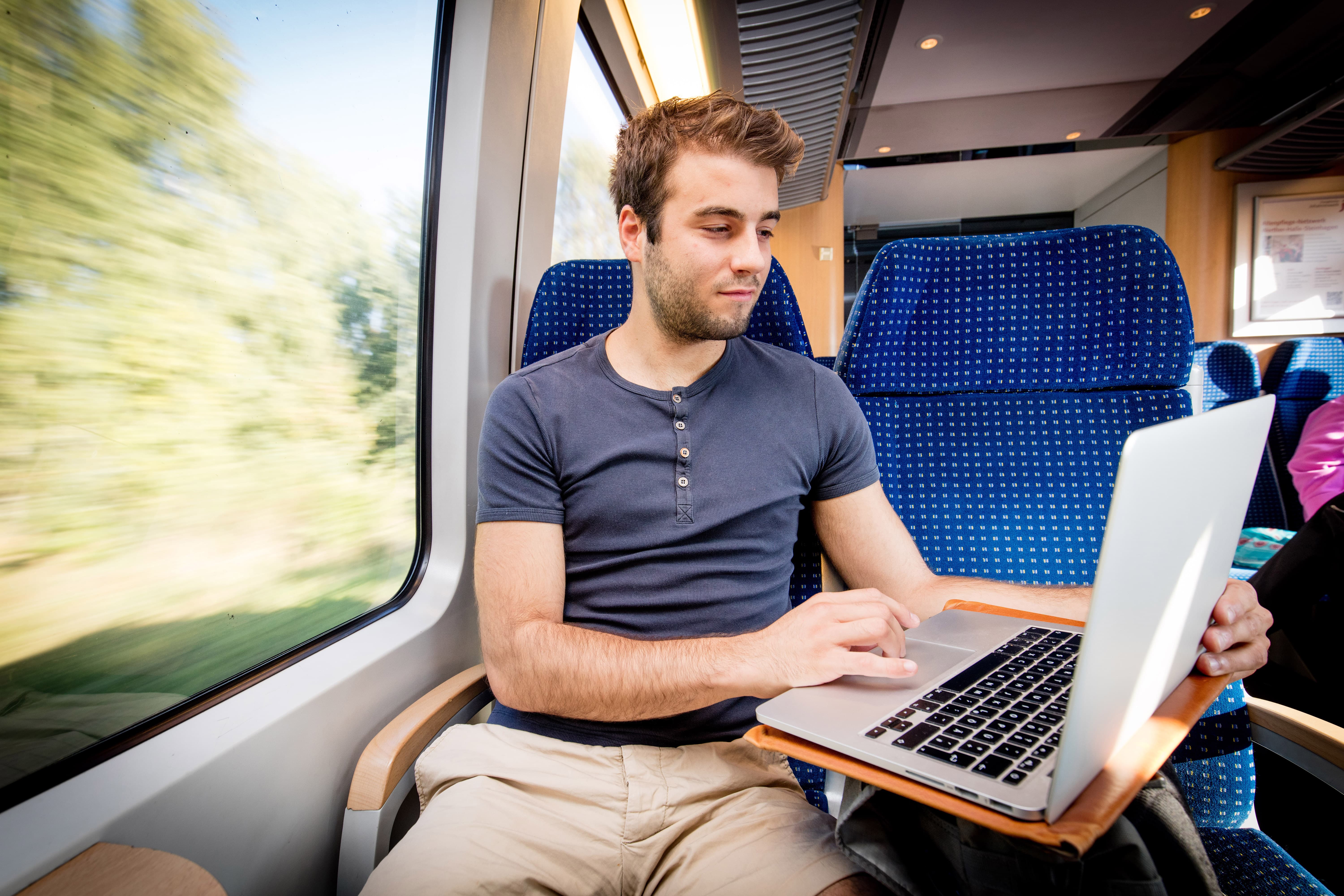 Junger, sommerlich angezogener Mann lädt im Nahverkehrszug Dateien auf seinem Laptop herunter.