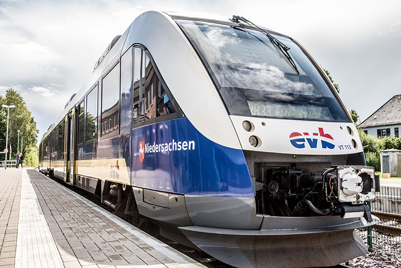 Blau-weiß-gelber Zug mit rot-weiß-blauem Logo der evb steht am leeren Bahnsteig